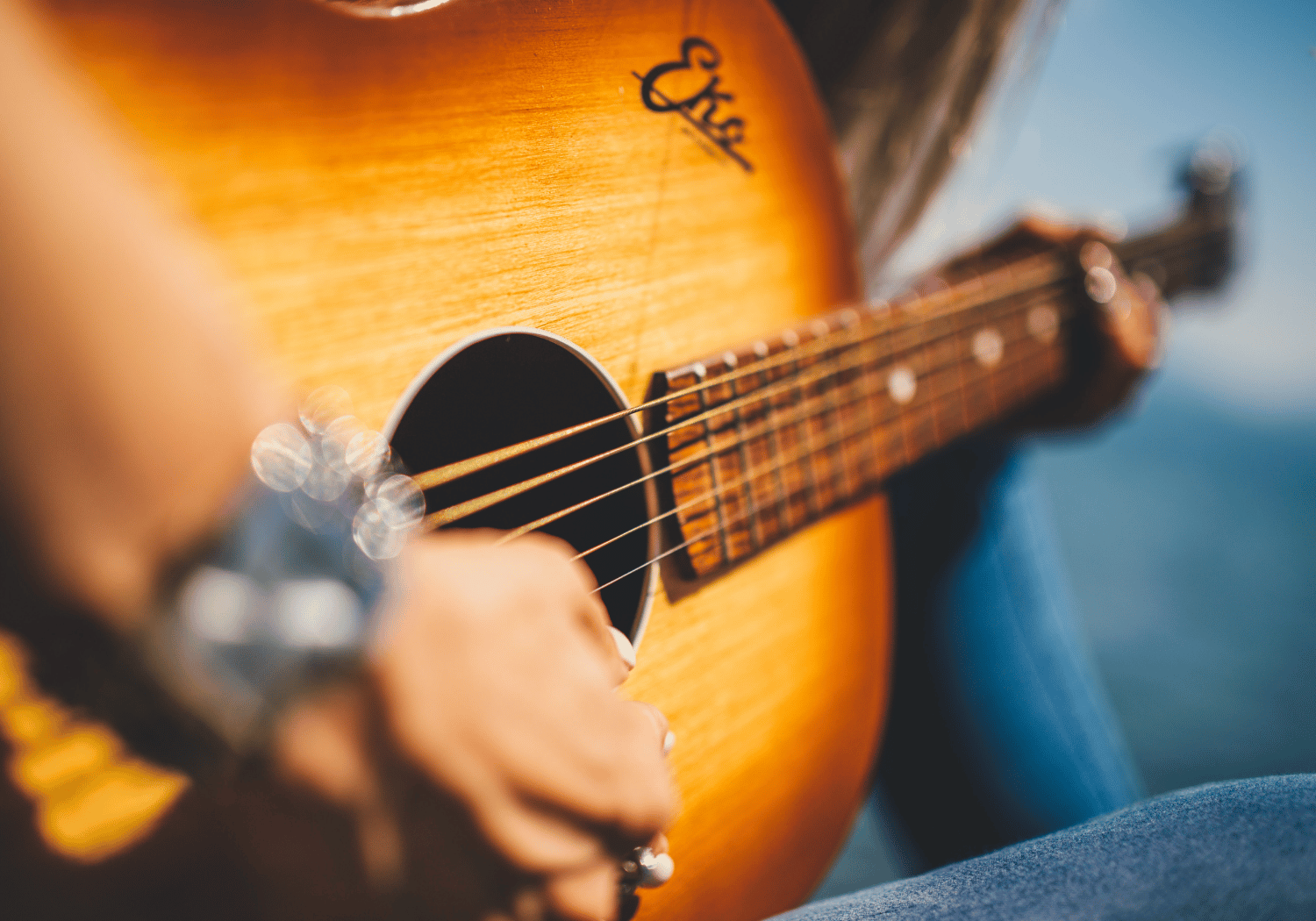 Close-up of a person playing a sunlit acoustic guitar by a body of water as they strum the strings.