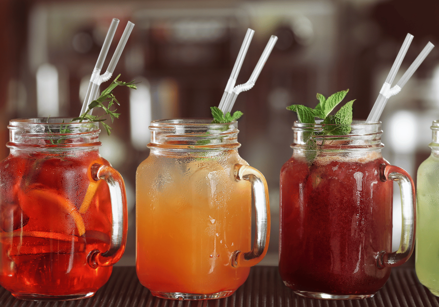Three mason jar drinks with ice and mint sprigs, each with a white striped straw, lined up on a bar counter (red, orange, and dark red beverages).