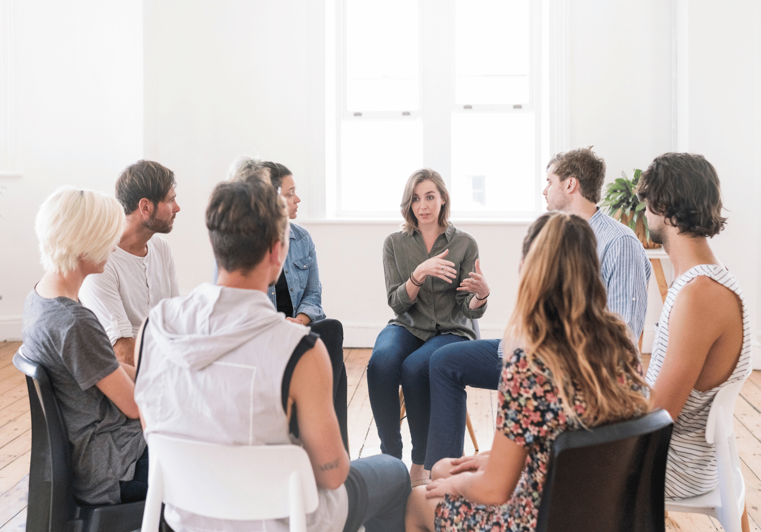 A woman in a green shirt leads a group discussion, sitting in a circle with others in a bright room.