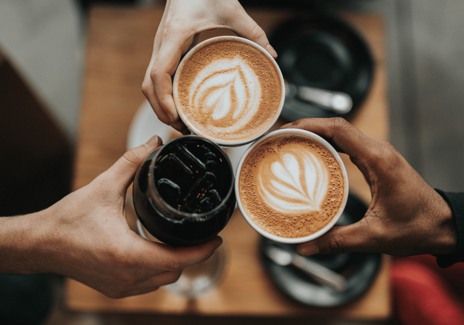 Three hands clink coffee cups in a toast, two with latte art rosettas and one glass of iced drink on a wooden table in a cozy setting