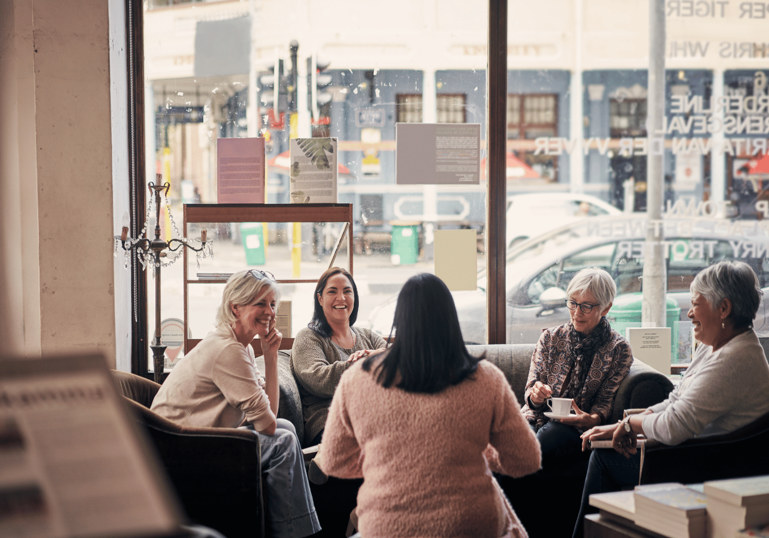 Five women sit together in a cafe, smiling and chatting by a large window with coffee cups on the table.